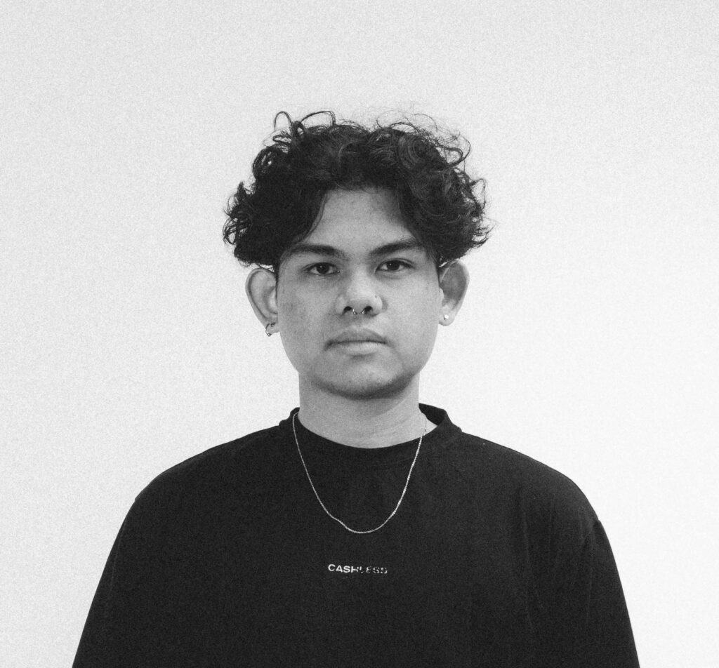 Black and white photo of Ryan Syach, CEO & Executive Producer at SNXP Studio, featuring a young man with curly hair, wearing a black t-shirt and a chain necklace, gazing thoughtfully at the camera.